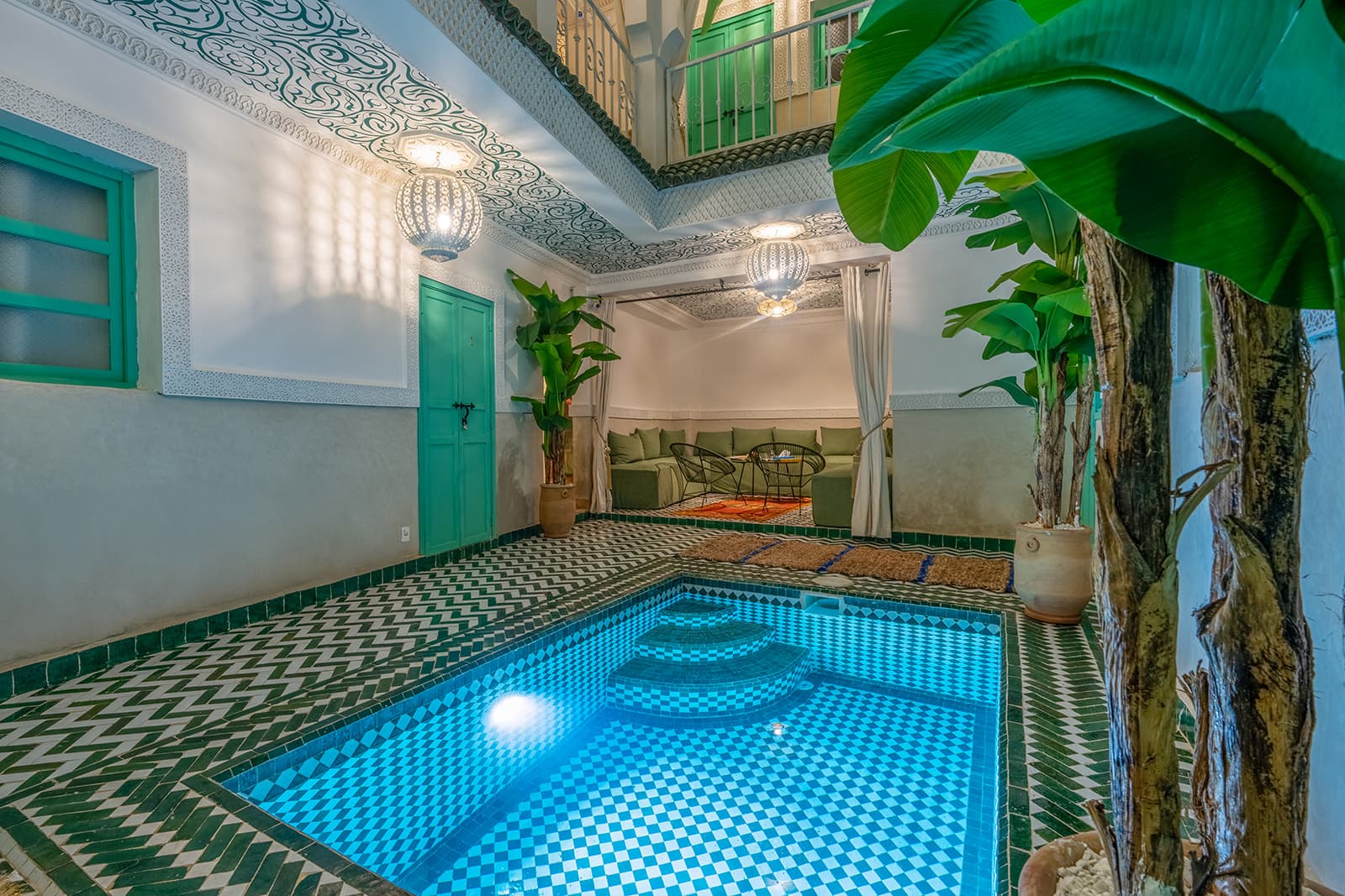 Le patio du riad : un bassin bordé de zellige vert, des bananiers, un plafond peint d'arabesques et des lanternes ciselées.