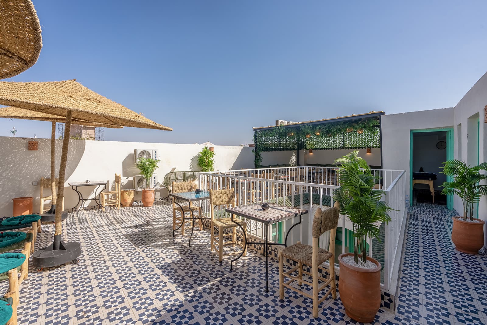 La terrasse en plein jour : sol de carreaux de ciment bleus et blancs, parasols de paille et jarres de terre cuite le long des murs à la chaux.