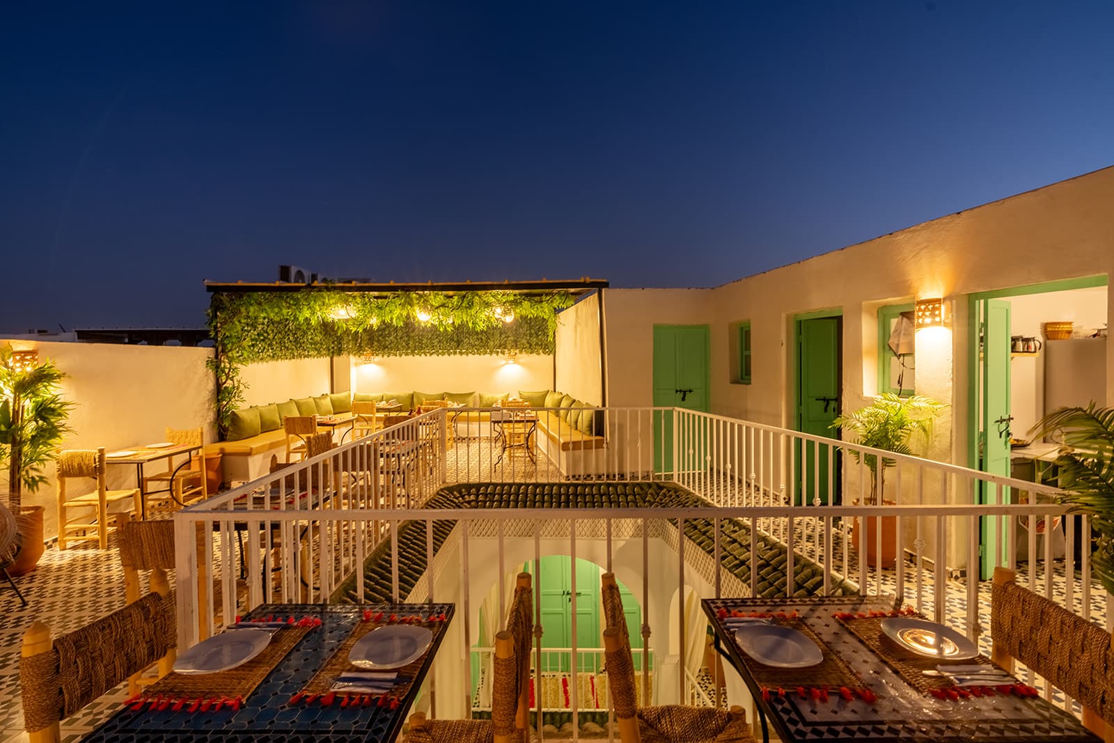 La terrasse la nuit : tables dressées, pergola tapissée de feuillage éclairé, portes vert menthe et appliques chaudes le long des murs.