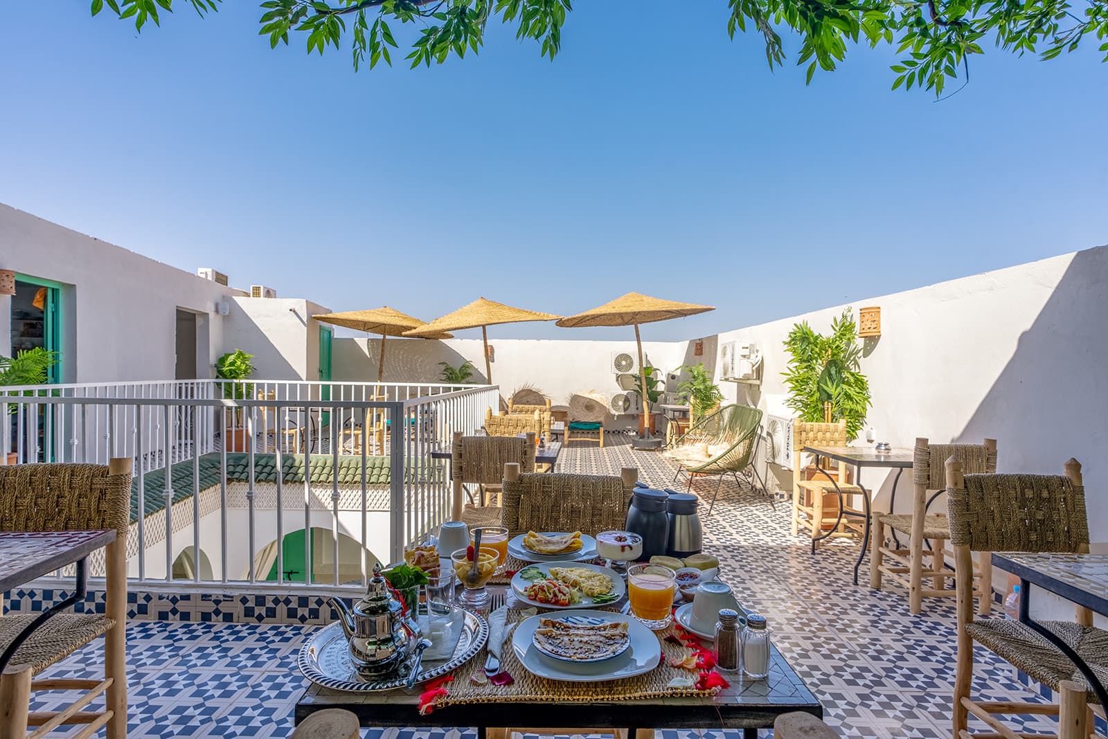 Une table dressée pour le petit-déjeuner sur la terrasse, théière en argent et jus pressés, sous un ciel bleu profond et des parasols de paille.